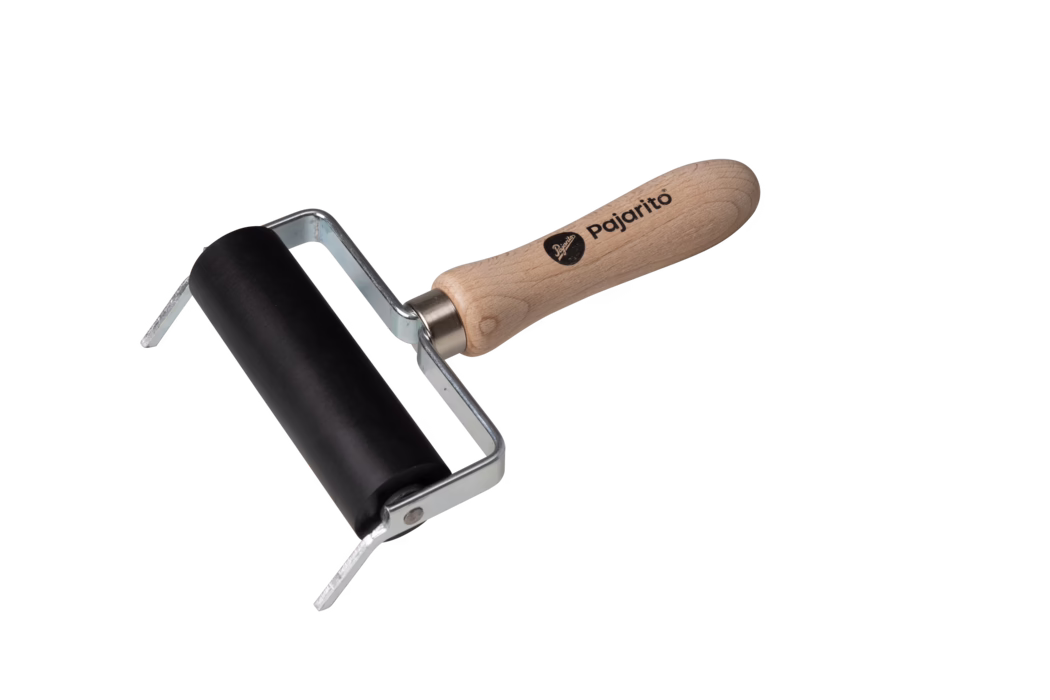 Pajarito_Dummy_Pressure roller support_036300_2021-02 Ein kleiner Handrakel (Brayer) vor weißem Hintergrund. Er besitzt eine glatte schwarze Gummiwalze in einem gebogenen silbernen Metallgestell und einen ergonomisch geformten hellen Holzgriff mit dem Markenaufdruck "Pajarito". Das Metallgestell befestigt die Walze mit sichtbaren Nieten; der Holzgriff zeigt natürliche Holzmaserung und ein abgerundetes Ende. Das Werkzeug dient zum gleichmäßigen Verteilen von Druckfarbe oder Lack auf Druckplatten, Linoleum oder Papier und wirkt sauber sowie unbenutzt. Die Komposition ist leicht diagonal, die Walze unten links, der Griff erstreckt sich nach oben rechts.