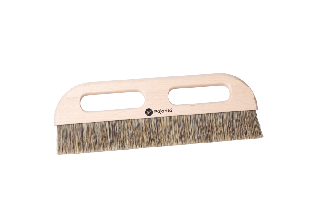 Nahaufnahme eines Holzhandbesens mit einem hellen Buchenholzgriff und dichten Naturborsten. Der flache rechteckige Griff besitzt zwei längliche ovale Handöffnungen für einen sicheren Zweihandgriff und ein kleines schwarzes Pajarito-Logo in der Mitte. Die Borsten bestehen aus gemischten beige- und braunfarbenen Fasern, sind gleichmäßig auf eine gerade Kante geschnitten und eignen sich zum Kehren von feinem Staub und kleinen Schmutzpartikeln auf Böden oder Arbeitsflächen. Das Modell ist auf einem sauberen weißen Hintergrund fotografiert und zeigt die glatt geschliffene Holzmaserung, abgerundete Ecken und das praktische ergonomische Design.