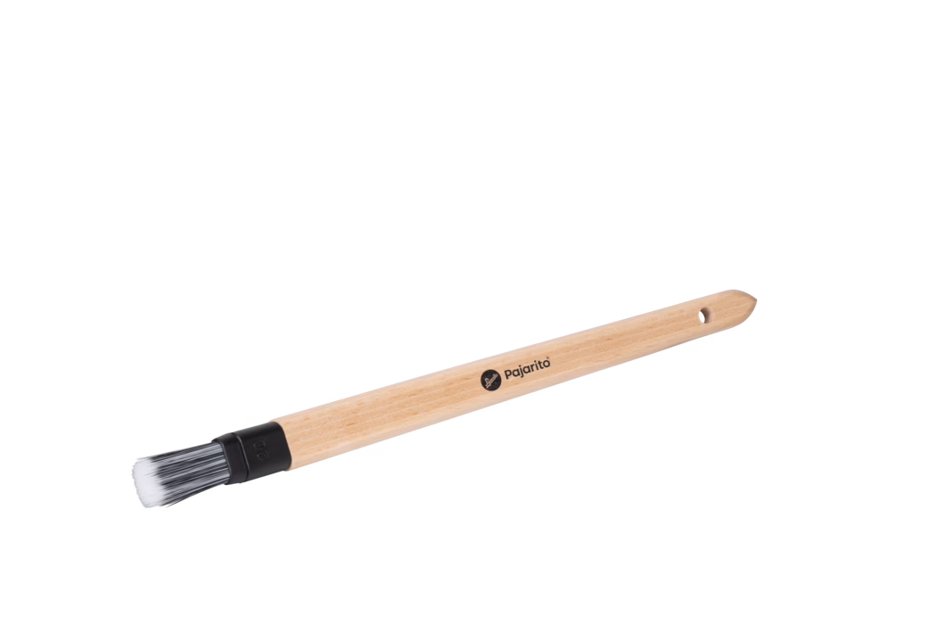 Ein kleiner Malpinsel mit einem schlanken Griff aus naturbelassenem Holz, einer schwarzen Kunststoffzwinge und kurzen synthetischen Borsten, die an den Spitzen weiß und an der Basis dunkler sind. Auf dem Griff befindet sich ein kleines schwarzes ‚pajarito‘-Logo sowie ein eingebohrtes Aufhängeloch am Ende. Der Pinsel liegt diagonal auf weißem Hintergrund und ist von oben fotografiert.